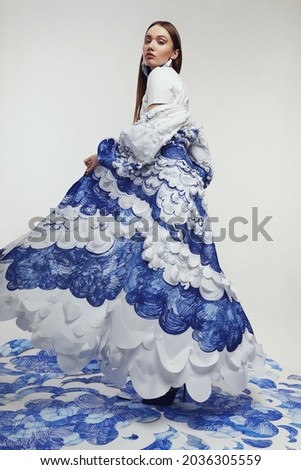 Woman in long fantastic cape with blue and white waves. Drawing and ruffles on clothes. Model standing on the blue waves drawing                                