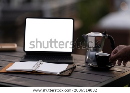 Photo of a white blank screen computer tablet putting on a wooden table surrounded by steel kettle, notebook, and coffee cup over the natural outdoors as a background.