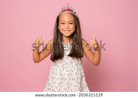 adorable happy smiling little child girl in crown princess celebrating on pink background with thumbs up. birthday party.