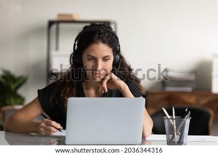 Focused female adult student in headphones using laptop, attending online lesson, virtual class, listening webinar, watching training, writing notes, making video call. Remote learning on internet Royalty-Free Stock Photo #2036344316