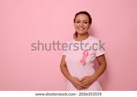 A smiling mixed race woman in pink T-shirt with pink satin ribbon symbolizing International Breast Cancer Day, expressing solidarity and support for breast cancer patients and survivors. 1 st october Royalty-Free Stock Photo #2036353559