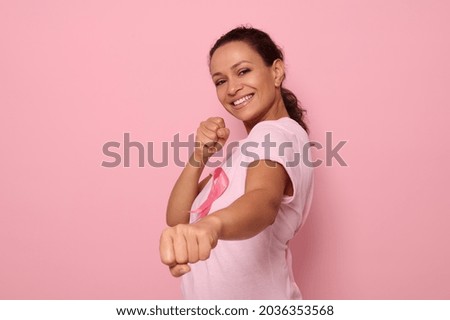 Pretty woman in pink t-shirt and cancer awareness ribbon stands in a fighting stance to mark the fight against cancer, in honor of October 1, smiles looking at camera, colored background, copy space Royalty-Free Stock Photo #2036353568