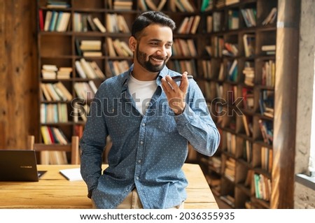 Smiling Indian man using convert voice into text mobile app, holding smartphone, talking into a mic. Bearded hispanic guy recording and sending voice message, writing sms by voice standing in library