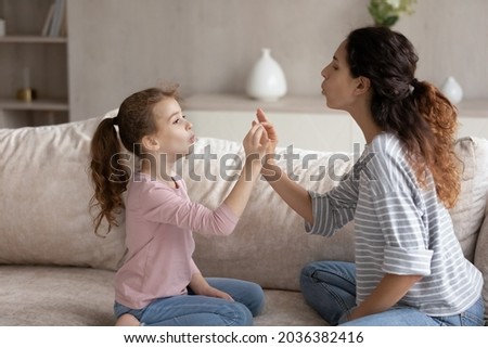 Caring young hispanic mum teaching small preschool child daughter pronouncing words correctly. Adorable cute girl doing vocal exercises with professional teacher, sitting on cozy couch at home. Royalty-Free Stock Photo #2036382416