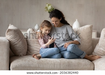 Happy small kid girl using telephone applications, playing mobile games or spending time online resting on comfortable sofa with affectionate caring young mother, modern technology addiction concept.
