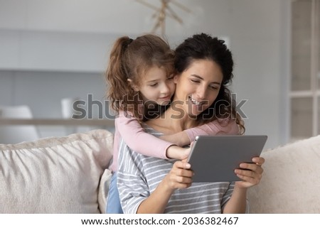 Loving adorable child daughter cuddling smiling hispanic mother or nanny, using digital computer tablet applications, choosing goods shopping in internet store, playing games, spending time online.