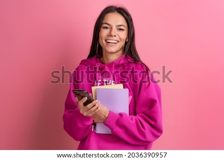 hispanic pretty woman in pink hoodie smiling holding holding notebooks and using smartphone posing on pink background isolated