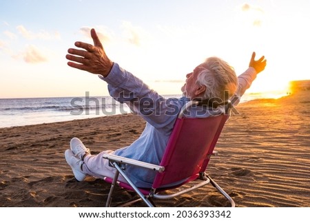 One mature and old man enjoying the summer vacations alone at the beach sitting in a small chair looking to the sea. Male person feeling free with opened arms. Freedom concept. Royalty-Free Stock Photo #2036393342