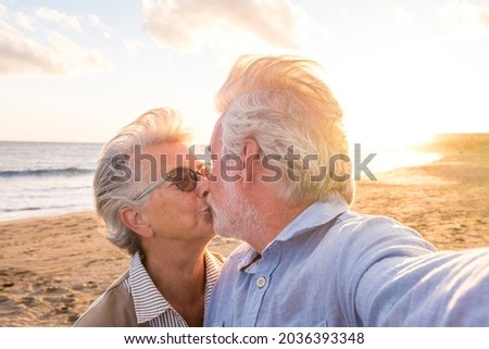 Portrait of couple of mature and old people enjoying summer at the beach looking to the camera taking a selfie together with the sunset at the background. Two active seniors traveling outdoors.