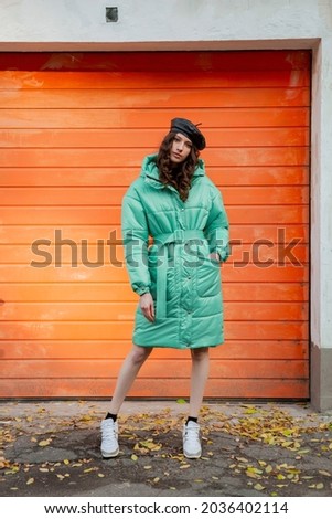 stylish woman posing in winter autumn fashion trend puffer coat and hat beret against orange wall in street wearing sneakers