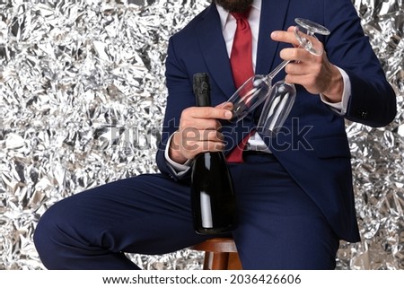 elegant businessman in navy blue suit recommending champagne bottle, holding glasses and posing in front of tinfoil background in studio