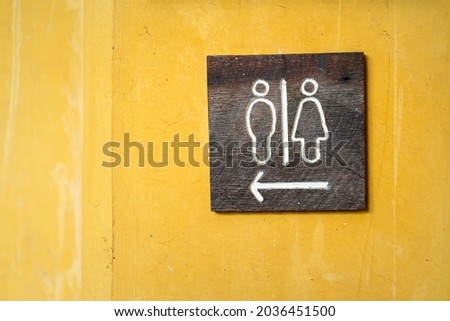 Toilet direction information wooden board with gender icon and arrow which is installed on yellow wall. Sign object photo. Close-up and selective focus.
