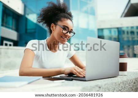 African American College Girl Using Laptop Learning Online And Browsing Internet Doing Homework In Modern University Outdoors. Student Typing On Computer, Studying Wearing Eyeglasses. Side View
