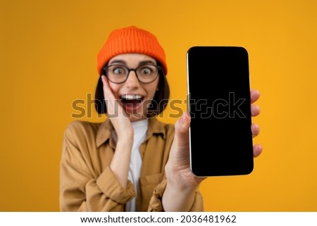 Wow, amazing useful app. Surprised young lady holding smartphone with black blank screen in hand, showing device, closeup. Gadget with empty free space for mockup template