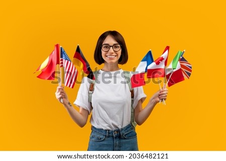 Happy lady showing bunch of diverse flags cheerfully smiling at camera over yellow studio background, excited female student recommending foreign language studying school Royalty-Free Stock Photo #2036482121