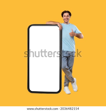 I Recommend. Full body length of cheerful young man leaning on large smartphone with white blank screen, showing thumbs up sign gesture. Gadget with empty free space for mock up, great mobile offer