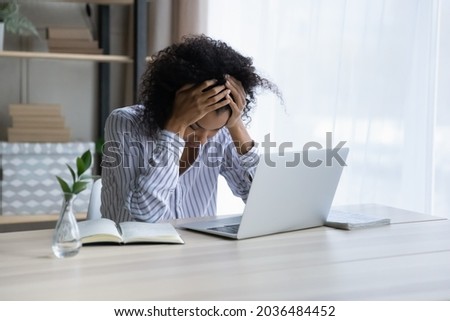 Professional burnout. Depressed upset mixed race female sit at workplace by computer hug head feel unmotivated sick lack of sleep. Desperate young black woman having problems at work missed deadline Royalty-Free Stock Photo #2036484452