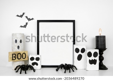 Halloween mock up. Black frame on a white shelf with rustic wood ghost decor, spiders and black candle. Portrait frame against a white wall with bats. Copy space.