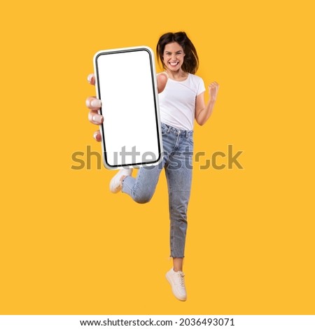 Yes. Full body length portrait of joyful woman jumping up, making winner gesture with clenched fist and showing cellphone with free empty space for mobile app on white screen, orange studio background