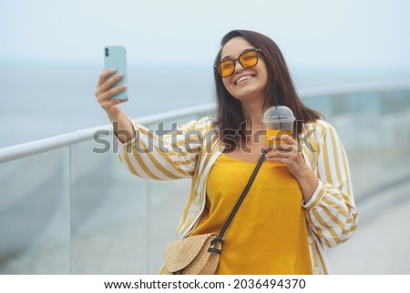 Cheerful and happy young overweight brunette woman 30-35 years old makes a selfie on a mobile phone. Fashionable plus size girl in casual clothes takes pictures of herself on a smartphone on vacation