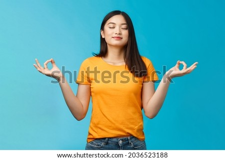 Peaceful charming relaxed asian girl buddhist meditating, breathing, inhale fresh air practice yoga, close eyes smiling relieved, stand lotus nirvana pose, reach zen, standing blue background Royalty-Free Stock Photo #2036523188