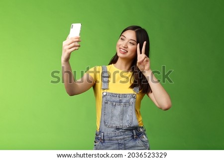 Tender cute asian brunette female, hold smartphone, posing selfie, look telephone screen, photographing, show victory peace sign, joyfully chatting friend abroad, stand green background