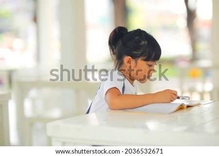 Little kid girl asian wearing a jeans jumpsuit reading the books on the desk with a magnifying glass. Which increases the development and enhances outside the classroom learning skills concept.