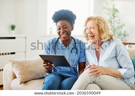 Doctor or nurse caregiver showing a tablet screen to  senior woman and laughing at home or nursing home