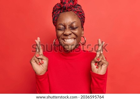 Happy dark skinned woman crosses fingers for good luck smiles broadly dreams to fullfil something she wanted believes in fotune wears scarf on head and turtleneck isolated over red background Royalty-Free Stock Photo #2036595764