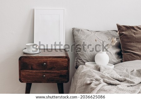 Vertical white picture frame mockup. Retro wooden bedside table. Modern white ceramic vase with dry Lagurus ovatus grass and cup of coffee. Beige linen, velvet pillows in Scandinavian bedroom. 