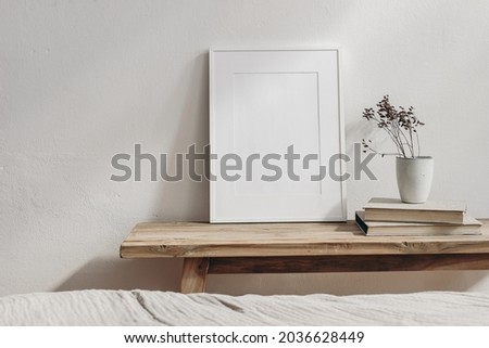 Vertical white picture frame mockup. Vintage wooden bench, table. Cup with dry grass on pile of books. White wall background. Scandinavian interior, neutral color palette. Selective focus. Art display