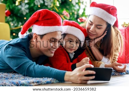 Happy Asian family father mother and daughter wears sweater with red and white Santa Claus hat lay down on carpet floor using smartphone camera taking selfie photo celebrating Christmas eve together.