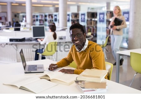 Portrait of smiling african-american male student with laptop and book in public library. High quality photo