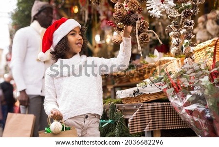 Smiling girl with toy in hand choosing decorations at Christmas market, father on background