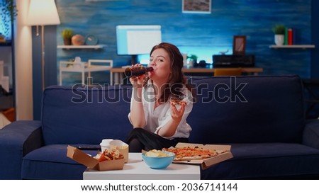Person eating slice of pizza on couch and looking at camera in living room. Caucasian woman with fast food meal from delivery at home while watching television and laughing after work