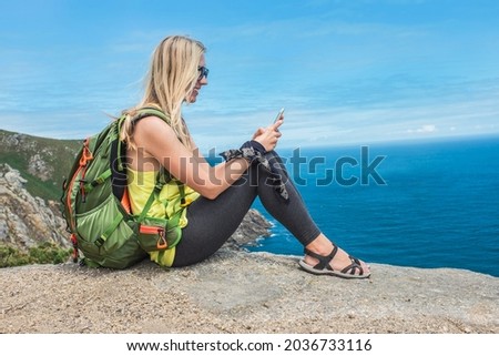 Woman hiker, hiking backpacker traveler camper with her phone camera on the top of mountain in sunny day under sun light. Beautiful sea landscape view.