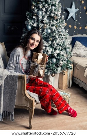 Young beautiful brunette woman in pajamas unpacks a New Year's gift under a Christmas tree