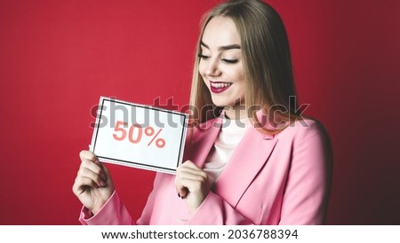 Beautiful Caucasian Blonde Woman With Long Hair Smiling and Happy Holding a Sign That Says 50%, Wearing a Bright Pink Blazer and Red Lipstick on a Red Colored Background.