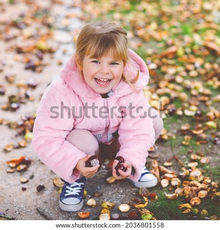 Adorable cute toddler girl picking chestnuts in a park on autumn day. Happy child having fun with searching chestnut and foliage. Autumnal activities with children
