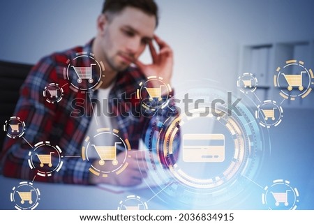 Businessman wearing red shirt is taking notes in front of laptop with interface with icons of credit card and cart. Office workplace with laptop in background. Concept of online buying and purchasing