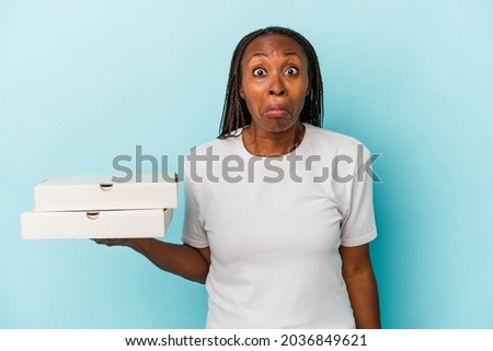 Young african american woman holding pizzas isolated on blue background shrugs shoulders and open eyes confused.