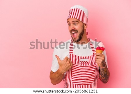 Young caucasian ice maker man with tattoos holding ice cream isolated on pink background  points with thumb finger away, laughing and carefree.