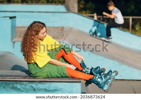Young redhaired woman in green and yellow clothes and orange stockings with curly hairstyle put on rollerblades in skate roller park