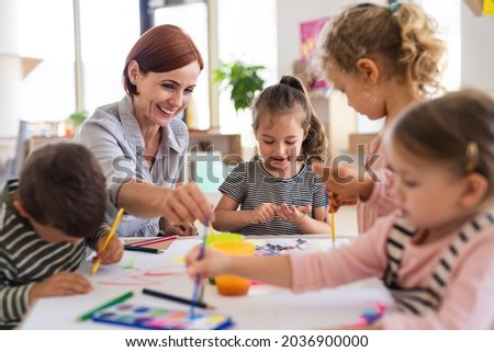 Group of small nursery school children with teacher indoors in classroom, painting. Royalty-Free Stock Photo #2036900000