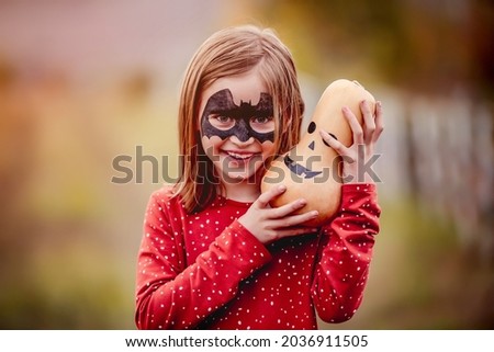 halloween portrait of smiling little girl with bat mask makeup holding big pumpkin above head outdoors