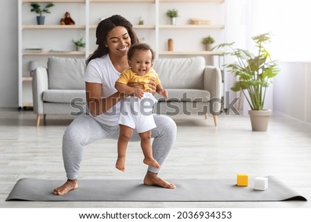 Weight Loss After Pregnancy. Happy Black Mom Training At Home With Infant Baby, Making Squats While Holding Her Cute Toddler Son In Hands, Exercising Pilates On Fitness Mat In Living Room, Copy Space Royalty-Free Stock Photo #2036934353