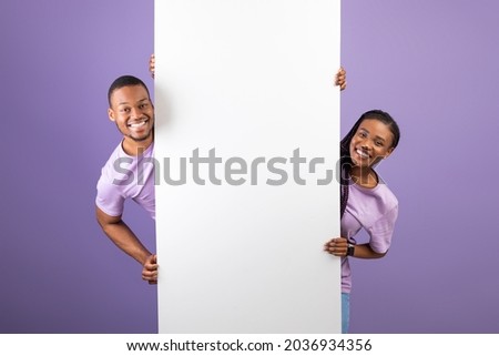 Smiling young black couple holding white vertical advertisement board, demonstrating free copy space for your text or design, positive guy and lady peeking out banner, purple violet background Royalty-Free Stock Photo #2036934356