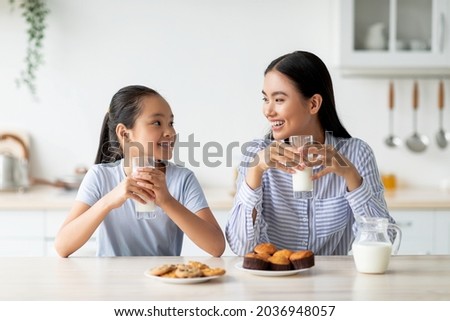 Pretty asian teen girl drinking milk with her mom and eating cookies, sitting at kitchen table, having pleasure conversation and smiling to each other, copy space