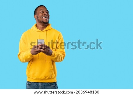 Joyful African Man Using Cellphone Texting And Looking Aside At Copy Space Posing Standing Over Blue Background, Studio Shot. Great New Mobile Application For Phone Concept Royalty-Free Stock Photo #2036948180