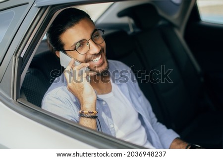 Cheerful young indian man sitting in taxi back seat, talking on cellphone with friend and smiling, shot from window, copy space. Handsome long-haired arab guy using transportation service Royalty-Free Stock Photo #2036948237